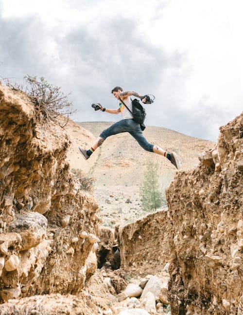 Full body side view of risky male hiker jumping over stony formations during trip in nature with hilly area