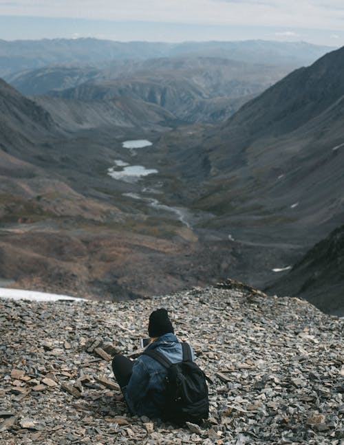 Back view of anonymous hiker with backpack sitting on edge of stony mountain above valley with creek during trip in highlands