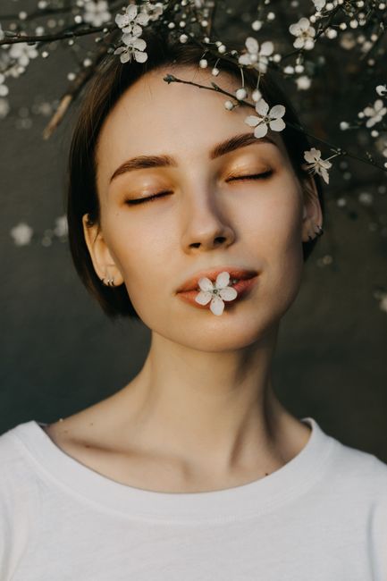 Tranquil young female with short hair and closed eyes with small white flower in mouth standing near blooming branches on gray background