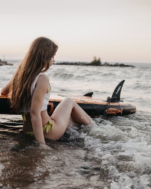 Side view of young female in swimwear with SUP paddleboard sitting on sandy beach near waving sea in summer