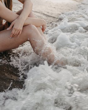 Crop unrecognizable slim female sitting on sandy beach near waving sea water on summer evening