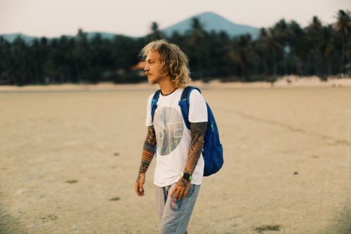Free Young male with tattoos and blue backpack in casual clothes walking on sandy beach near green trees while looking away on summer day Stock Photo