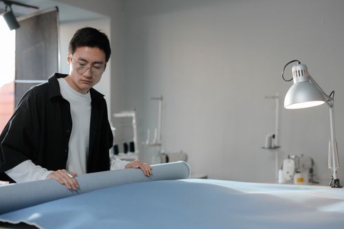 Free Fashion designer examining a blue fabric roll in a well-lit workspace. Stock Photo