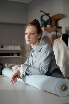 A thoughtful fashion designer leans on a fabric roll in her studio, ready for work.