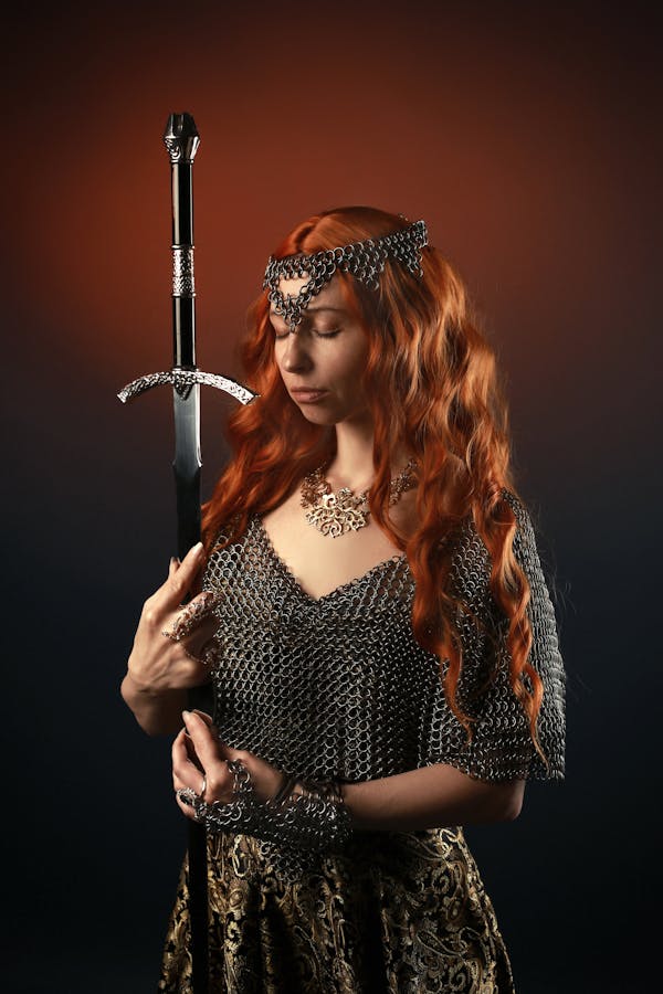 Concentrated young female with ginger long hair with necklace and wearing chainmail armor and costume of warrior with sword standing with closed eyes