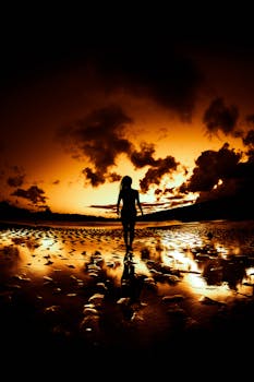 A dramatic silhouette of a person walking on a reflective beach shore during a stunning sunset.