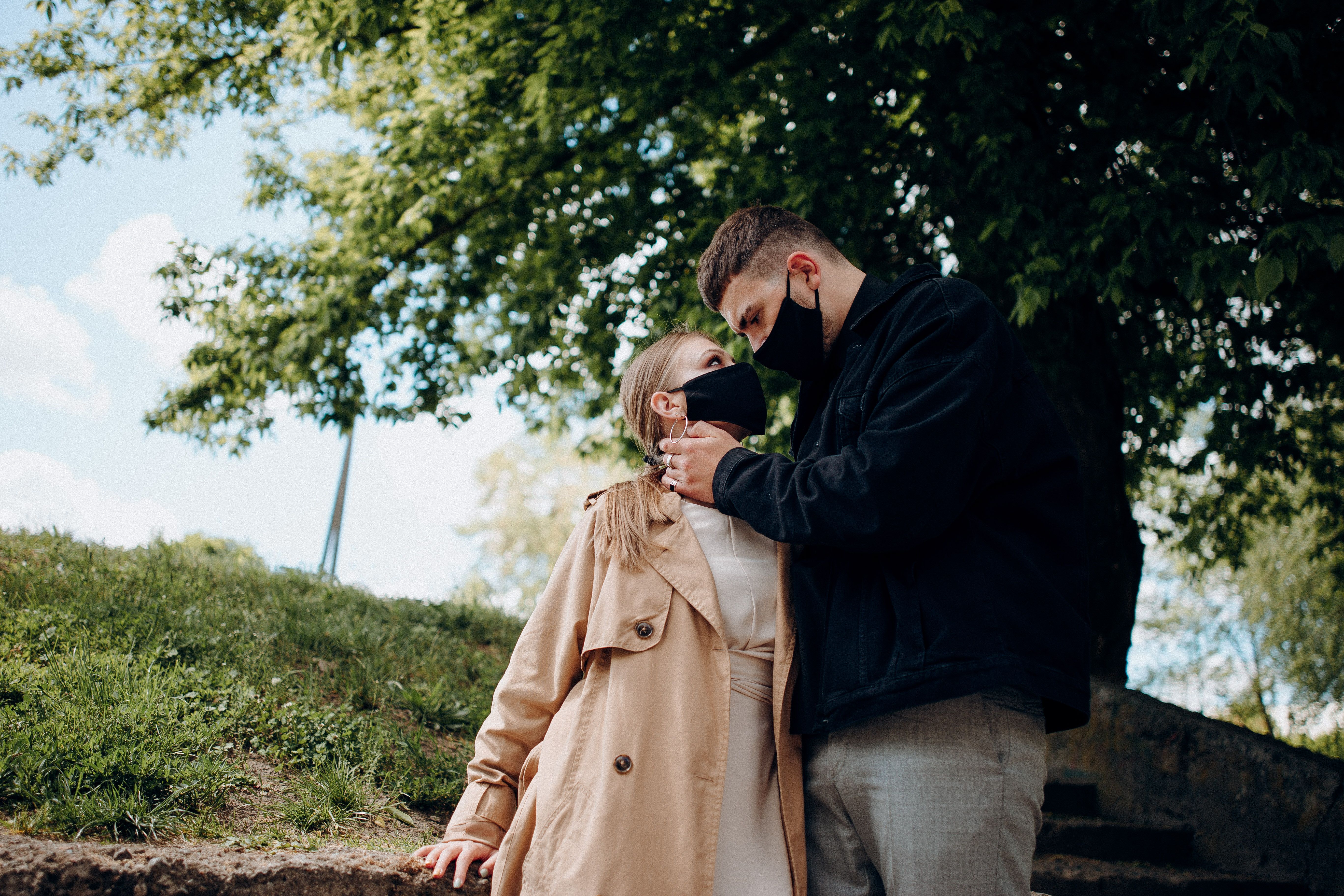 Young couple in stylish clothes and masks hugging and looking at each other while standing in park near tree in autumn