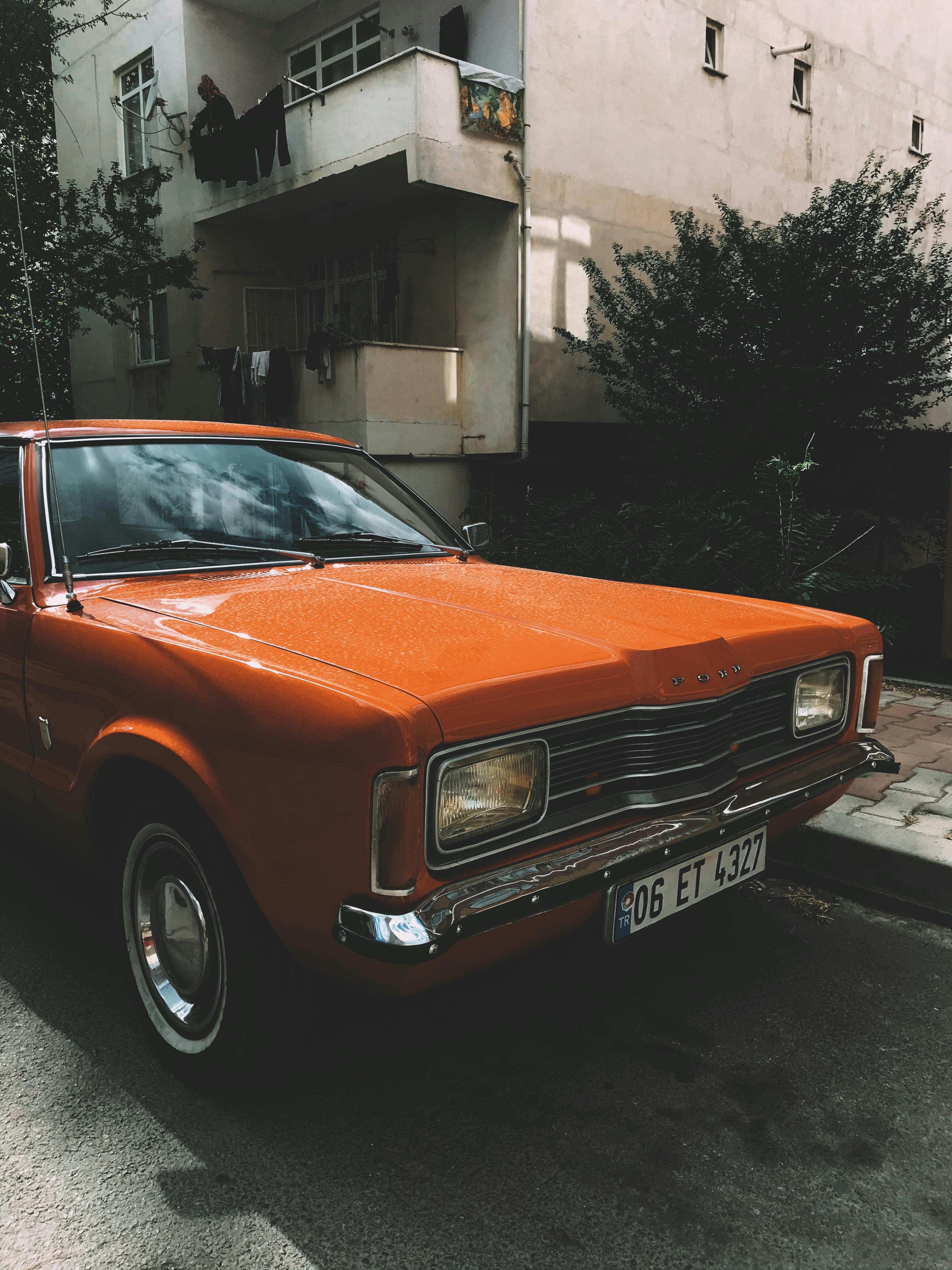 Old timer red automobile parked on sidewalk near tall residential house with balconies on street with green trees in town