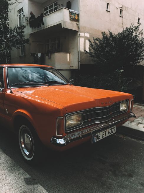 Old timer red automobile parked on sidewalk near tall residential house with balconies on street with green trees in town