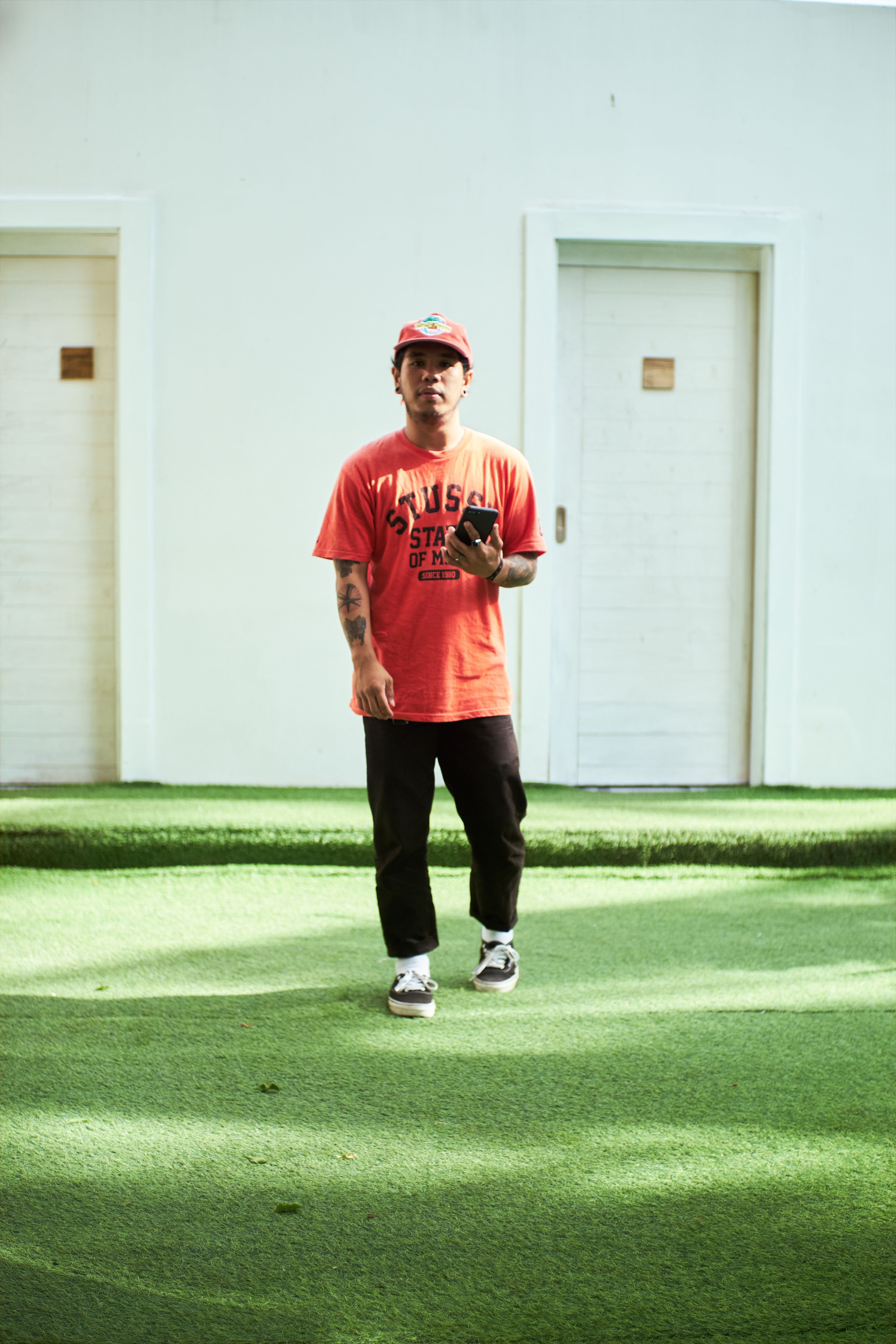 A man in an orange shirt with tattoos walks on grass, holding a cellphone.