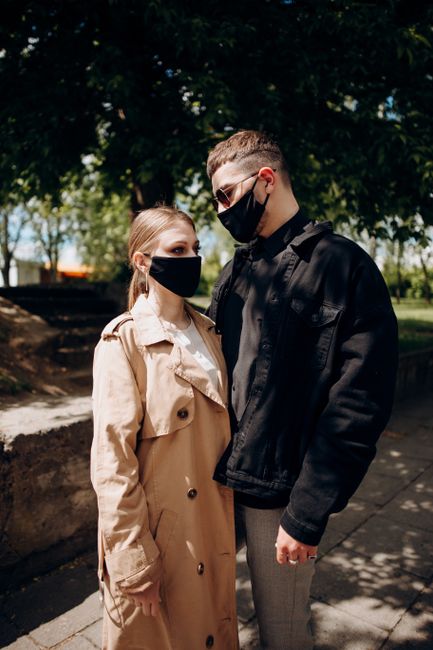 Couple in trendy outerwear and black masks standing on pavement in park on sunny autumn day