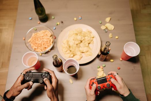 Two gamers playing video games with snacks and drinks on table, a perfect setup for gaming night.