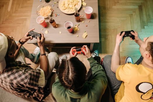Three friends play video games with snacks and drinks on a table in an indoor setting.