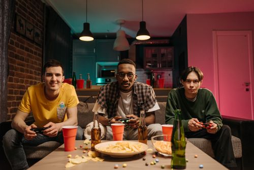 Free Three young men playing video games in a cozy living room with snacks and drinks. Stock Photo