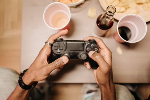 A person holding a gaming controller surrounded by snacks and drinks on a wooden table.