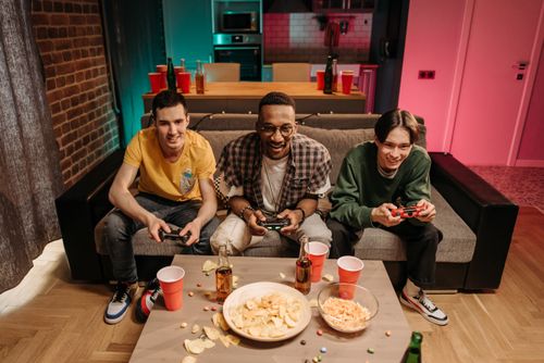 Free Three friends enjoying a fun gaming session on a sofa with snacks and drinks around. Stock Photo