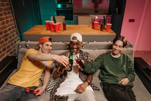Free Three friends clink beer bottles while relaxing indoors, enjoying leisure time together. Stock Photo