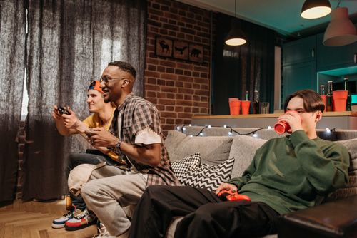 Free Three friends playing video games and drinking on a cozy couch, creating a lively indoor atmosphere. Stock Photo