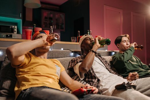 Three friends relaxing with drinks and gaming on a cozy indoor couch setting.