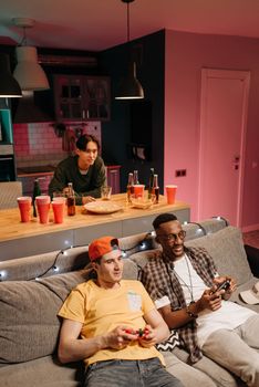 Three friends enjoying a video game party with drinks and gaming consoles in a cozy living room.