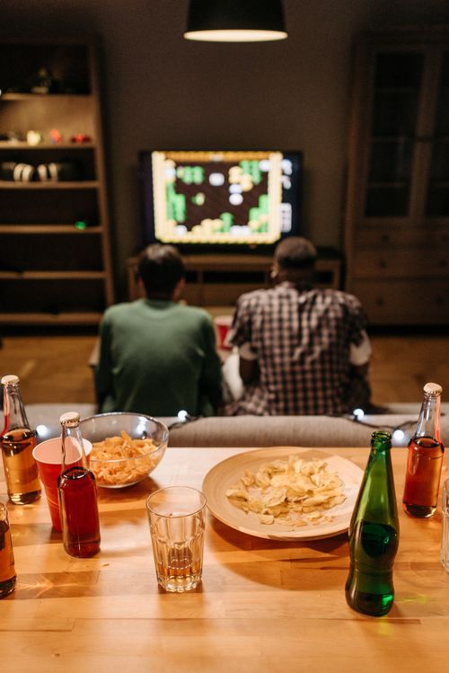 Free Two friends playing video games at home, surrounded by snacks and drinks on a table, enjoying leisure time. Stock Photo