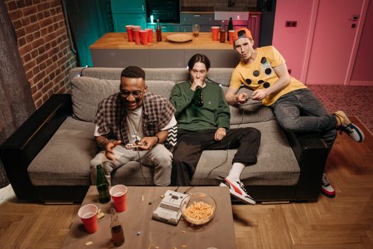 Three friends having fun gaming and drinking beer in a cozy indoor setting.