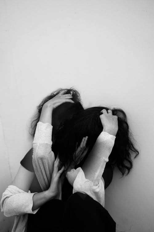 Black and white of faceless lady in casual clothes covering face with dark hair near white wall in daytime