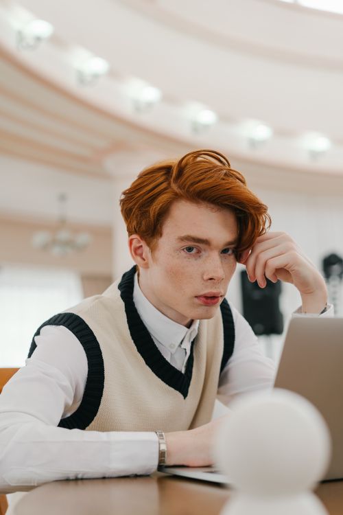 Free Thoughtful young man in a sweater vest working on a laptop inside a bright room. Stock Photo