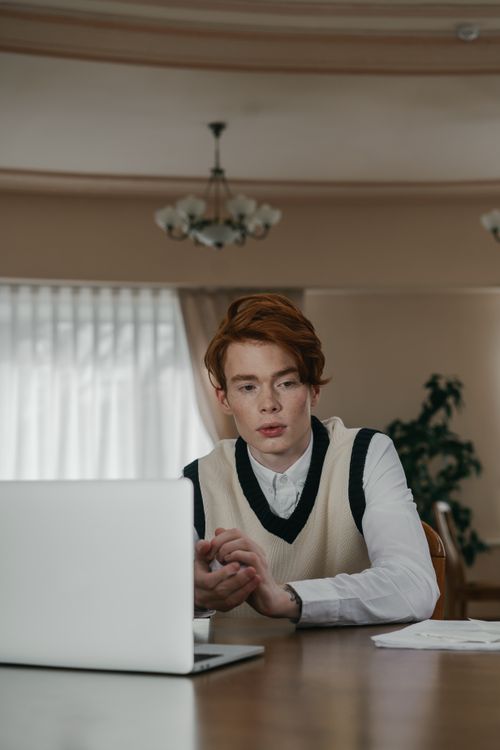 Free Young adult man in a sweater vest focused during an online meeting, seated indoors with natural light. Stock Photo