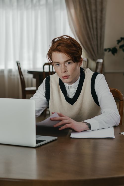 Free A young adult wearing a sweater vest focuses on online studies, seated at a table with a laptop. Stock Photo