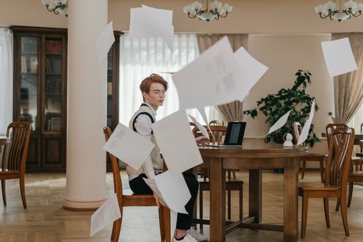 A young man looks upset in a library as papers fly around him, symbolizing academic stress.