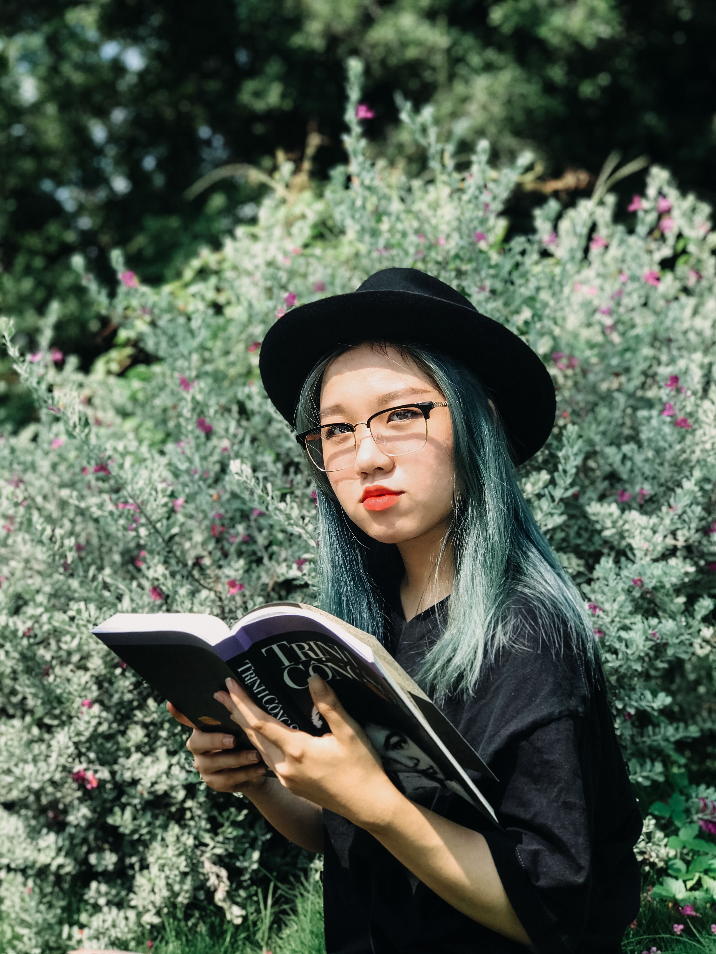 Young Asian woman with eyeglasses and hat reading book in a vibrant garden setting.
