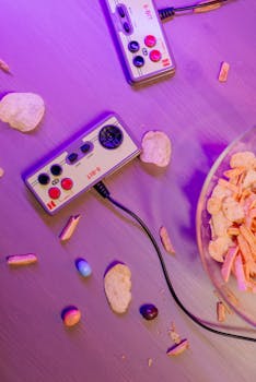 Retro video game controllers with scattered snacks on a table, illuminated by purple lighting.