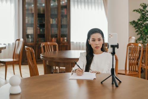 A Woman Holding a Pen Sitting at a Table with a Smartphone on a Tripod