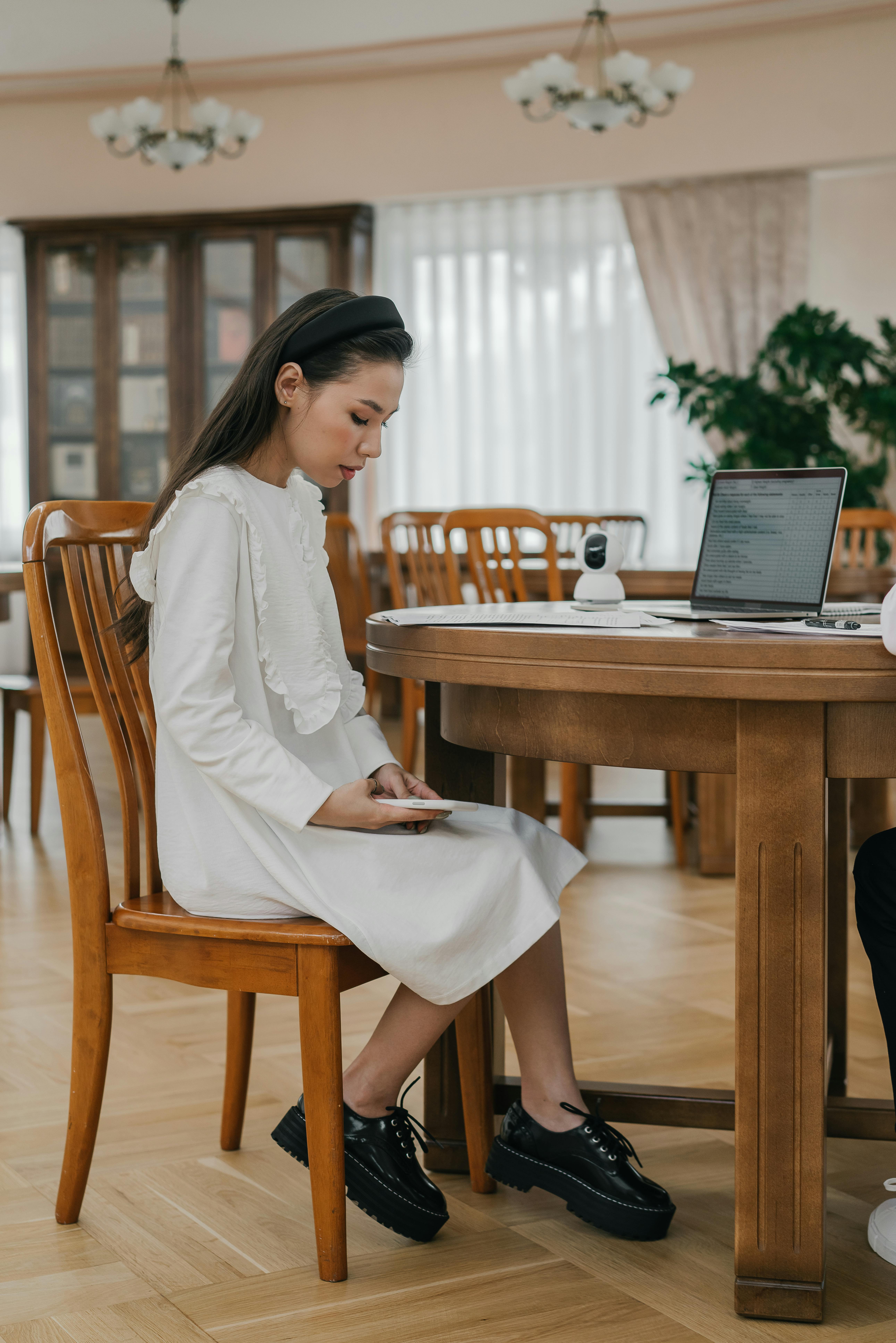 a woman in white dress sitting on a wooden chair while using her phone