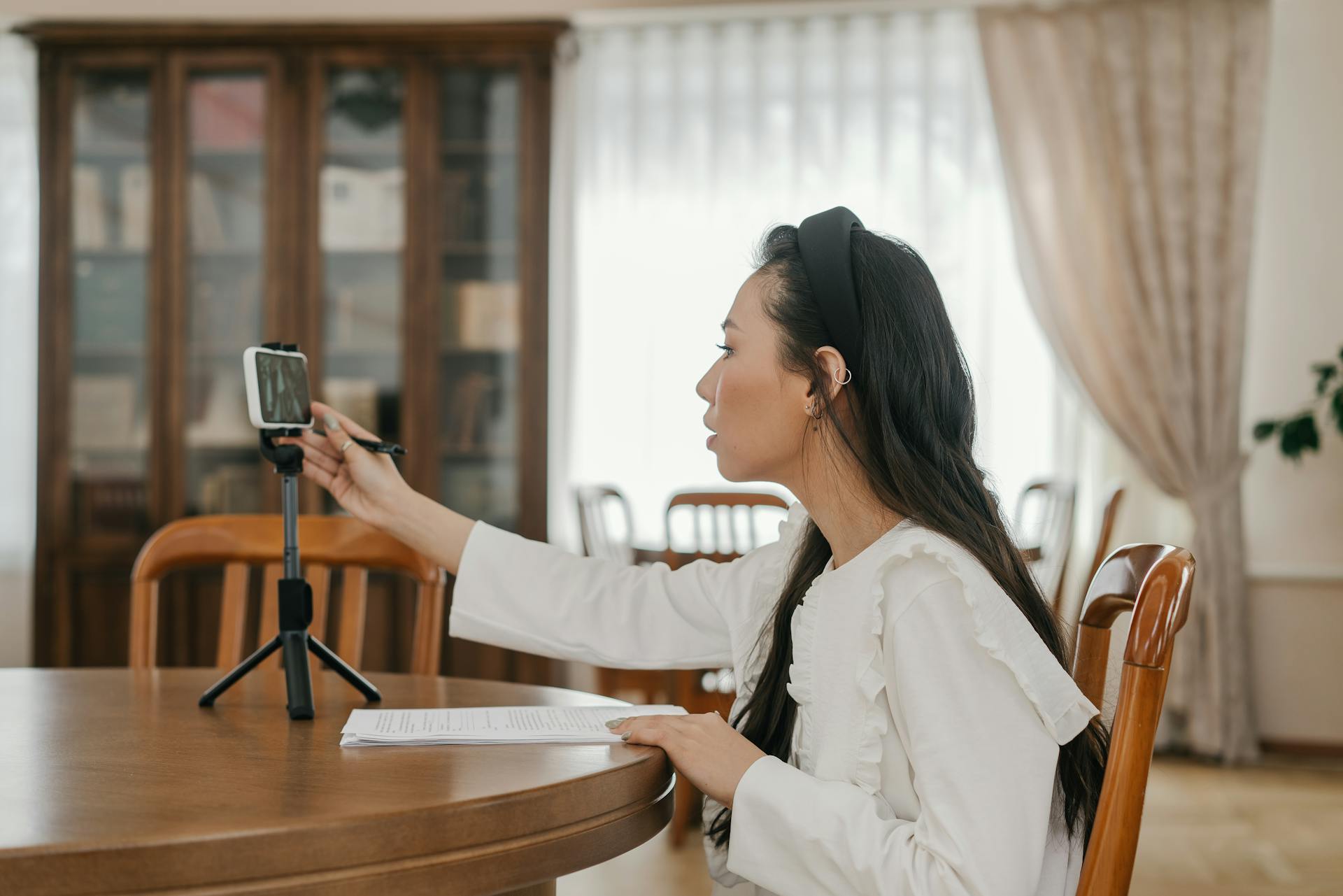 A Woman in White Long Sleeves Sitting on a Wooden Chair while Using Her Phone