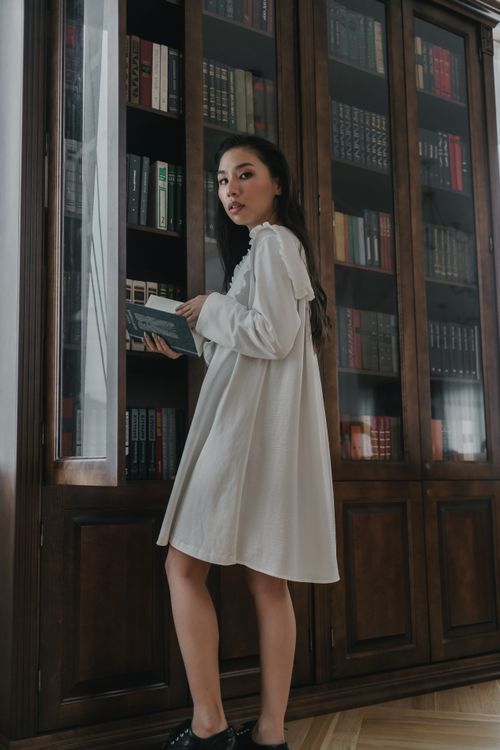Free A young woman in a white dress reads a book in a cozy home library setting. Stock Photo