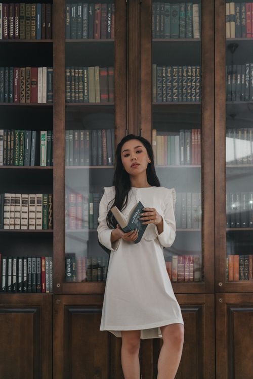 Free Asian woman in white dress holding books in a library setting. Stock Photo