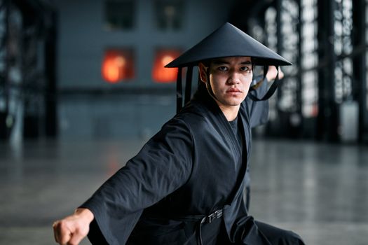 Martial artist wearing a black kimono and rice hat performing a striking pose indoors.