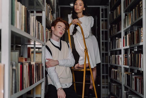 Woman in White Long Sleeve Dress Standing on Yellow Ladder beside Bookshelves