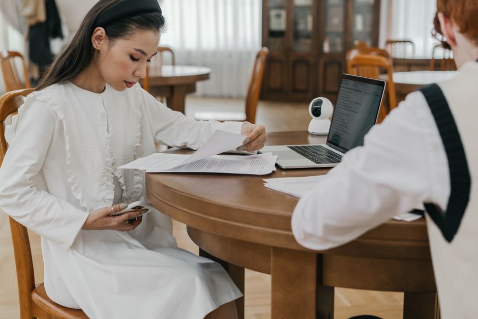 Two students focusing on their studies at a communal table, using laptops and documents. Two students focusing on their studies at a communal table, using laptops and documents.