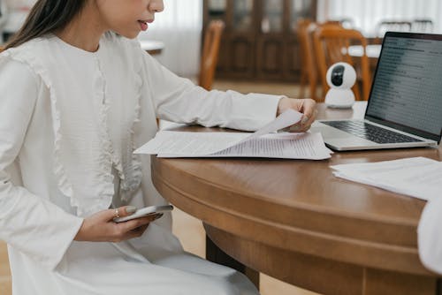 A Woman Sitting Near the Wooden Table while Holding Papers and Mobile Phone