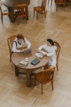 Two students working together at a round table in a library with books and laptops.