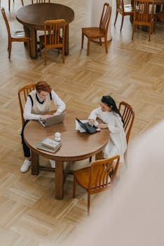 Two students engaged in studying with laptops at a wooden table indoors.