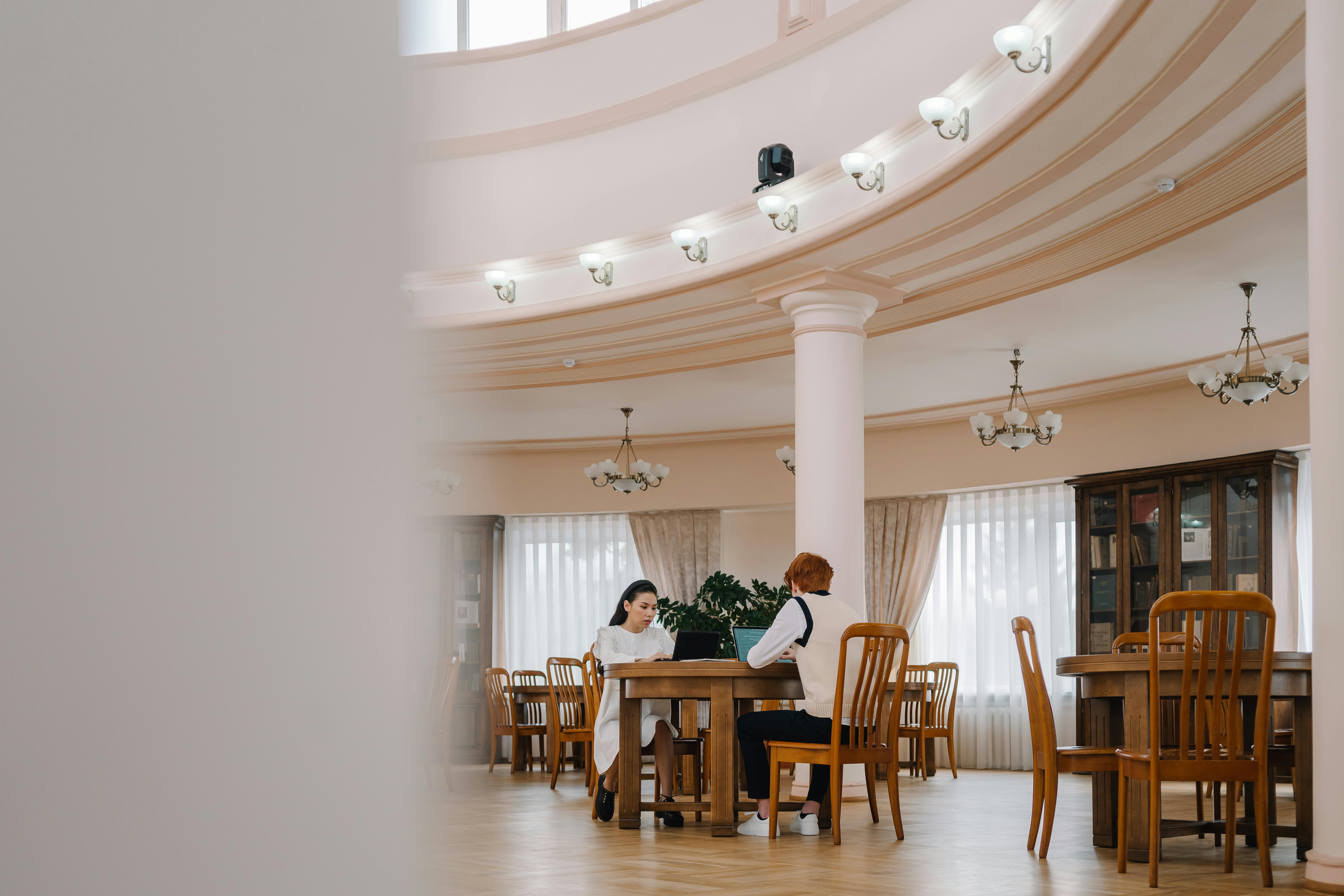 Two students studying at wooden tables in stately library with laptops and books.