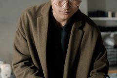 Asian man in coat with eyeglasses writing in a notebook indoors.