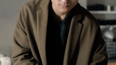 Asian man in coat with eyeglasses writing in a notebook indoors.
