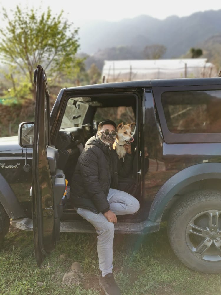 A Man Sitting On A Car With His Dog