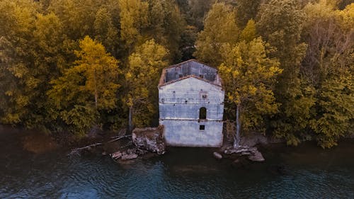 Drone view of old shabby ruined building located among autumn trees and shore of rippling river