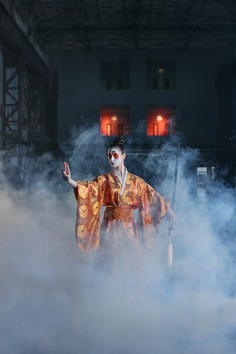 Performer In A Red Kimono With Golden Patterns And Geisha Makeup Posing With A Sword Among Clouds Of Smoke